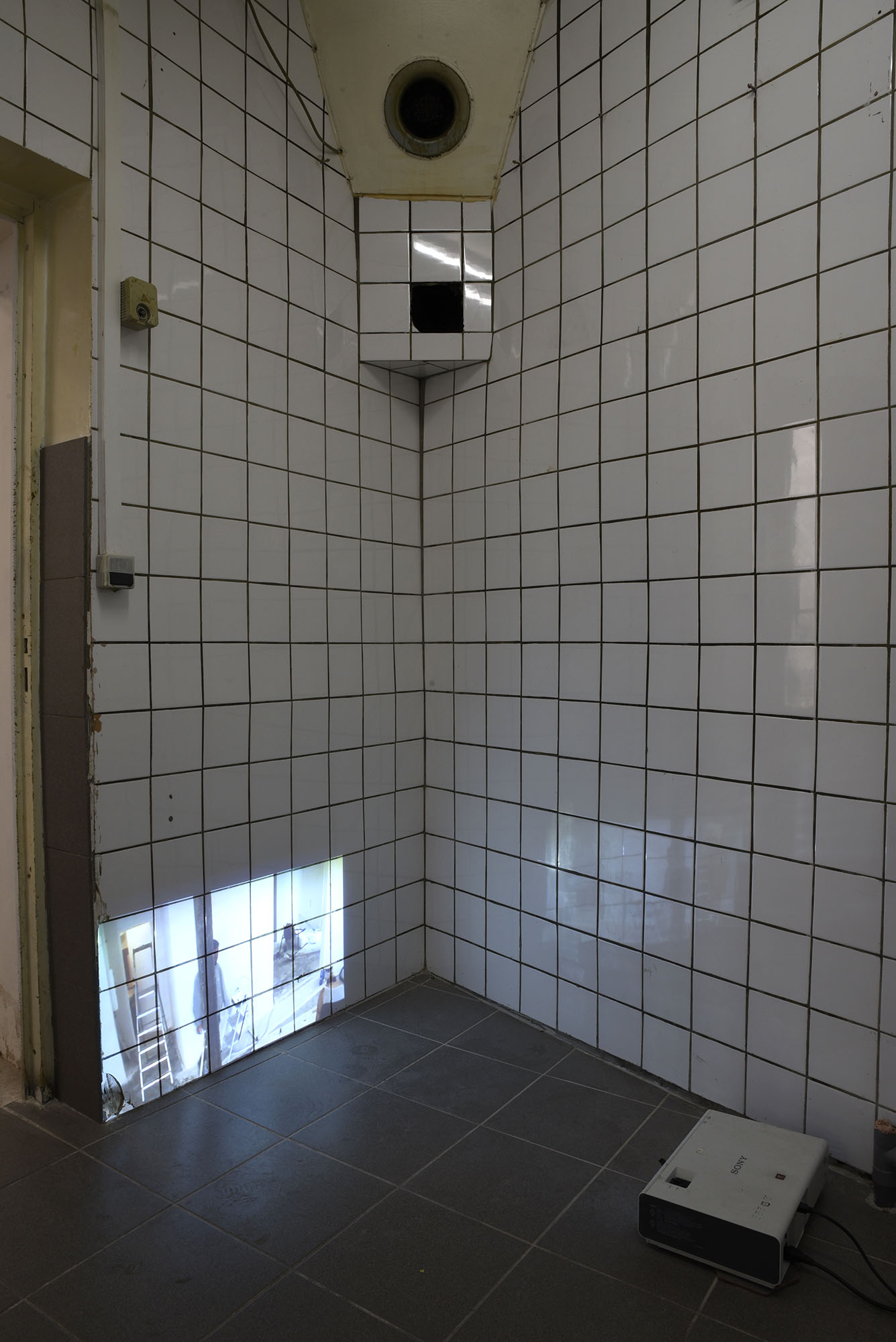 Dans un espace en carelage (mur et sol) on voit une image projeter sur le mur depuis un vidéoprojecteur posé à même le sol. Ce sont des gens en train de faire des travaux.